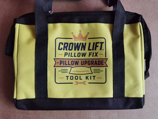 A yellow and black tool kit bag with a printed graphic that reads 'CROWN LIFT PILLOW FIX PILLOW UPGRADE TOOL KIT' with a crown and tool icon, photographed on a brown surface. Actual color of tool ag, yellow