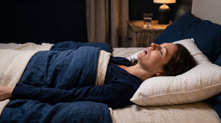 A woman lies on her back on a bed with white pillows and a navy blue blanket, in a dimly lit bedroom setting. Her mouth is open snoring, she has no head support