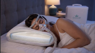 A woman sleeping on a bed with a nasal CPAP mask connected to a tube, using a crown lift insert in her pillow. A white Crown Lift  brand carrying case is visible in the background on the bed next to a bedside lamp.