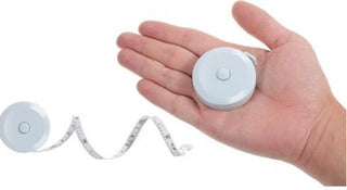 A white circular retractable measuring tape dispenser shown in a person's hand, with part of the white tape uncoiled on a plain white background. Used for measuring pillow height and head drop sink in pillow for use with crown lift upgrade tool kit.