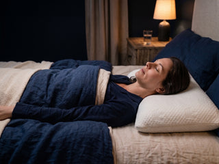 A woman lies asleep on her side on a bed with a white pocket pillowcase on her pillow with a crown lift inside navy blue bedding, and a bedside lamp glowing in the background.